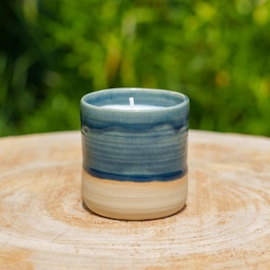 Può includere: Un portacandele in ceramica fatto a mano, blu e crema. Il portacandele cilindrico contiene una candela bianca con uno stoppino. Lo sfondo è verde sfocato, che suggerisce un ambiente esterno.