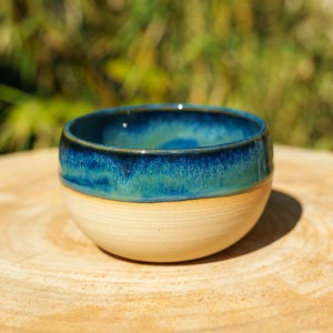 Könnte beinhalten: Eine kleine, runde Keramikschale mit einer glatten, unglasierten beigen Basis und einem glänzenden, tiefblauen und türkisfarbenen glasierten oberen Bereich. Das Innere der Schale ist mit derselben leuchtenden blau-türkisen Glasur versehen.