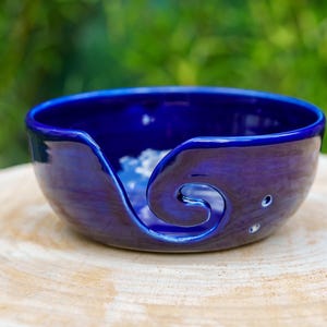 Peut inclure: Un bol à fil en céramique bleu foncé avec un motif en spirale et trois petits trous. Le bol est posé sur une surface en bois clair, avec un arrière-plan vert flou.