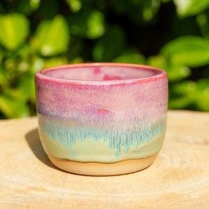 Peut inclure: Une petite tasse en céramique avec une forme arrondie et lisse dotée d'une finition émaillée réactive. L'extérieur présente un dégradé de couleurs, passant d'un rose vif au bord à une base bleu sarcelle et vert clair avec des coulures visibles.