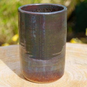 May include: A handmade ceramic tumbler with a dark, glossy finish. The cylindrical cup transitions from a deep brown base to a dark green and blue upper section. The interior is a darker brown. The cup is sitting on a wooden surface.