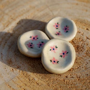 May include: Three small, off-white ceramic dishes with pink and blue floral designs. The dishes are arranged on a light brown, textured wooden surface. The dishes are round with slightly raised edges.