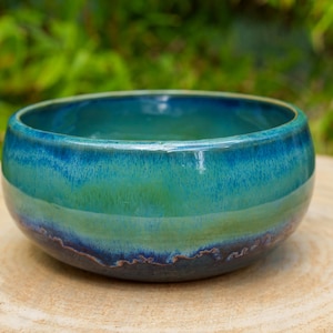 May include: A round ceramic bowl featuring a smooth, glossy glaze that transitions from a deep navy blue at the base to a vibrant turquoise and light green in the center, with a dark blue rim encircling the top edge of the vessel.