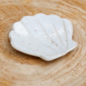 Puede incluir: Un plato blanco de cerámica con forma de concha y diseño moteado. El plato tiene un borde festoneado y está sobre una superficie de madera. Este plato decorativo es ideal para guardar objetos pequeños.