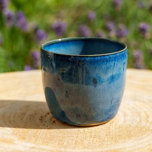 May include: A ceramic cup featuring a reactive glaze with deep navy blue and lighter sky blue tones. The vessel has a smooth, rounded silhouette with a slightly tapered base and a thin, dark rim, showcasing a glossy finish that reflects light.