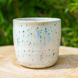 Peut inclure: Un gobelet en céramique de forme cylindrique avec une base légèrement effilée. L'extérieur présente un émail blanc brillant avec un motif moucheté de points bleus et de subtiles coulures beiges descendant verticalement du bord vers la base.