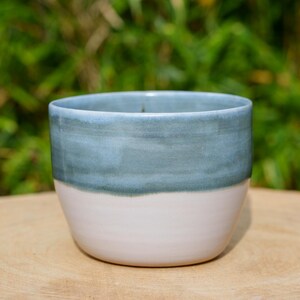Puede incluir: Taza de cerámica hecha a mano con un diseño de dos tonos. La mitad superior es de color verde azulado apagado, mientras que la mitad inferior es de un blanco suave. La taza tiene una forma redondeada y un acabado liso y esmaltado.