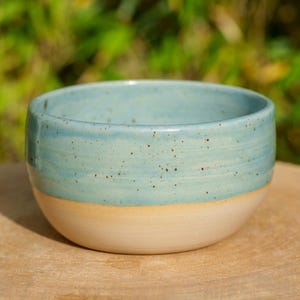 Peut inclure: Bol en céramique fait à la main, avec une glaçure bleu clair sur la moitié supérieure et une base crème. Le bol est de forme arrondie et parsemé de petits points bruns. Il est posé sur une surface en bois.