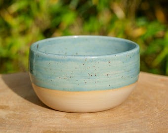 May include: A handmade ceramic bowl with a light blue glaze on the upper half and a cream-colored base. The bowl has a rounded shape and is speckled with small brown dots. The bowl is sitting on a wooden surface.