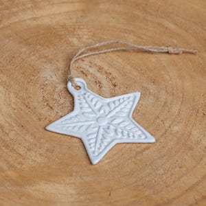 May include: A white star-shaped decoration with a detailed floral pattern, suspended by a natural twine string. The ornament is placed on a warm, wooden surface, creating a rustic look. Suitable for festive decorations.
