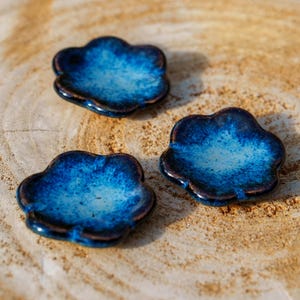 Peut inclure: Trois petites coupelles en céramique en forme de nuage, avec un glaçage bleu profond et brillant. Les bords ont une bordure plus foncée, presque noire. Les coupelles sont disposées sur une surface texturée brun clair, créant un contraste.