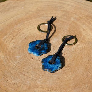 May include: Two keychains with blue ceramic flower-shaped pendants. Each pendant has a dark blue glaze with a hole in the center, attached to a black cord and a gold-colored ring. The keychains rest on a light brown wooden surface.