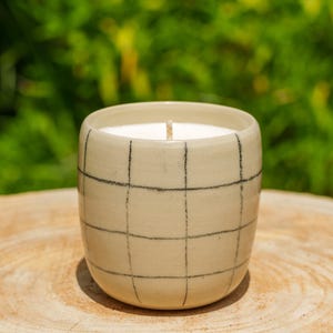 Peut inclure: Un récipient à bougie cylindrique en céramique de couleur crème claire avec un motif de grille noire peint à la main. Le contenant est rempli de cire blanche et comprend une mèche en coton centrée, présentant une finition lisse et brillante.