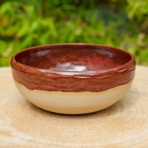 May include: A round ceramic bowl featuring a smooth, matte beige base and a glossy, textured rim in a deep reddish-brown glaze. The interior of the bowl is finished with a reflective, dark red glaze that contrasts with the light exterior.