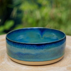 Op de afbeelding: Een ronde keramische kom met een diepblauwe en turquoise geglazuurde binnen- en buitenkant. Het glazuur heeft een gladde, glanzende afwerking met subtiele kleurverlopen. De onderkant van de kom is ongeglazuurd en toont de beige kleur van de klei.
