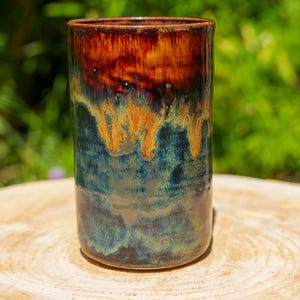 May include: A handmade ceramic tumbler with a unique glaze. The top is a deep reddish-brown, transitioning to orange and then to a teal-blue at the base. The mug sits on a wooden surface.