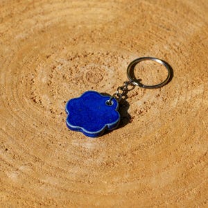 May include: A cobalt blue cloud-shaped keychain with a silver key ring. The keychain is made of a smooth, glossy material and has a white border. The background is a light brown, textured wooden surface.