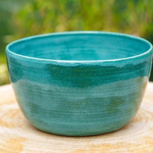Può includere: Una ciotola in ceramica turchese fatta a mano con finitura lucida. La ciotola ha una forma arrotondata e una superficie liscia e strutturata. La ciotola misura circa 15 cm di diametro e 10 cm di altezza. La ciotola è appoggiata su una superficie di legno.