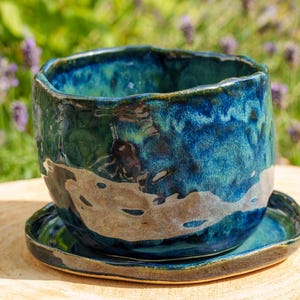 Peut inclure: Un pot en céramique avec une glaçure brillante bleu profond et sarcelle, doté d'un bord irrégulier et ondulé. La base du pot présente une section contrastante en terre cuite claire non émaillée. Il repose sur une soucoupe assortie en céramique.