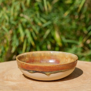 May include: A handmade ceramic bowl with a glossy finish. The bowl features a cream-colored base, transitioning to a brown and orange rim. The bowl is sitting on a wooden surface, with a blurred green background.