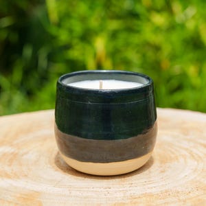Peut inclure: Une bougie artisanale dans un récipient en céramique. La bougie a une cire blanche et une mèche en bois. Le récipient est bleu foncé en haut, passant à une bande grise foncée et une base beige. La bougie repose sur une surface en bois.