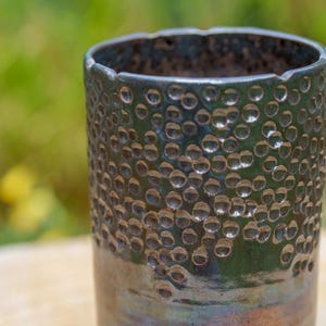Peut inclure: Un gobelet en céramique fait à la main avec une surface texturée de petits cercles en relief. Le gobelet a un glaçage bleu-vert foncé avec des reflets bruns et bleus, et un bord légèrement irrégulier. L'arrière-plan est flou, montrant un feuillage vert et jaune.