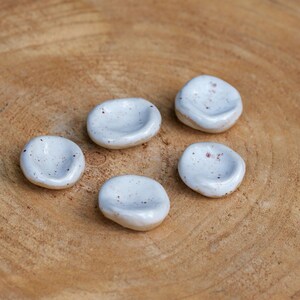 Peut inclure: Cinq petites coupelles en céramique blanche, avec un motif tacheté et une forme concave peu profonde. Les coupelles sont disposées sur une surface en bois brun clair avec des motifs de grain visibles. Les pièces en céramique ont une esthétique artisanale.