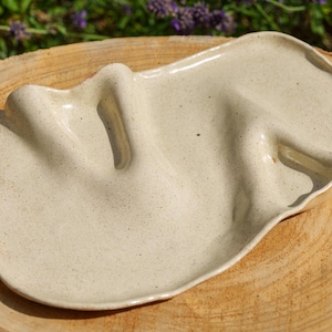 Peut inclure: Un plateau en céramique de couleur crème avec une finition mouchetée et une forme organique irrégulière. La surface présente deux crêtes distinctes, surélevées et ondulées, créant des indentations profondes, donnant un aspect fluide.
