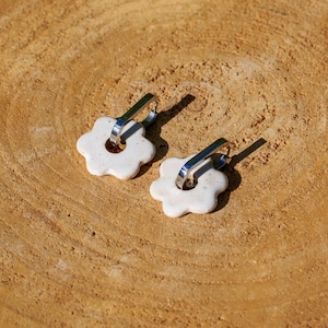 May include: Two white, flower-shaped earrings with silver-colored hooks. The earrings are set against a textured, light brown wooden surface, with shadows cast by the sunlight.