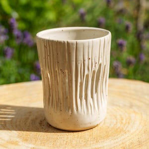 Peut inclure: Un gobelet cylindrique en céramique de couleur crème blanc cassé avec une finition mouchetée. La surface extérieure présente des rainures verticales sculptées et texturées qui descendent du bord vers la base, créant un motif linéaire.