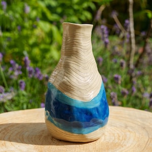 Op de afbeelding: Een keramische vaas met een smalle hals en een ronde basis. Het bovenste gedeelte heeft een lichtbeige kleur met horizontale gegraveerde lijnen. Het onderste gedeelte toont een levendig blauw glazuur met diverse blauwtinten.