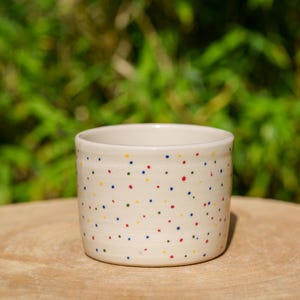 Könnte beinhalten: Eine kleine, cremefarbene Keramiktasse mit zylindrischer Form. Die Tasse ist mit einem Muster aus kleinen, gleichmäßig verteilten Punkten in Rot, Blau, Gelb und Grün verziert. Die Tasse steht auf einer Holzoberfläche, mit einem verschwommenen grünen Hintergrund.