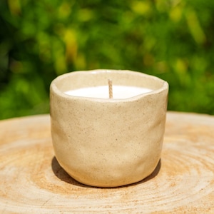 Peut inclure: Une bougie en céramique beige dans un récipient arrondi et texturé. La bougie a une cire blanche et une seule mèche centrée. La bougie repose sur une surface en bois brun clair, avec un arrière-plan vert flou.