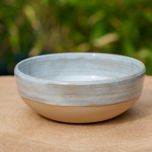 Puede incluir: Un pequeño cuenco de cerámica blanca con un borde marrón claro. El cuenco está sobre una superficie de madera.