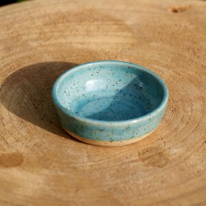 May include: A small, handmade ceramic bowl with a speckled turquoise glaze. The bowl has a rounded shape and a light-colored base. The bowl is sitting on a wooden surface.