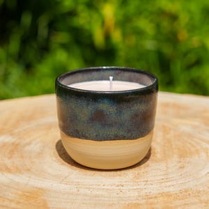 Op de afbeelding: Een keramische kaars in een donkerblauwe en bruingeglazuurde container. De kaars heeft een witte was en een enkele lont. De container staat op een lichtbruin houten oppervlak, met een vage groene achtergrond.