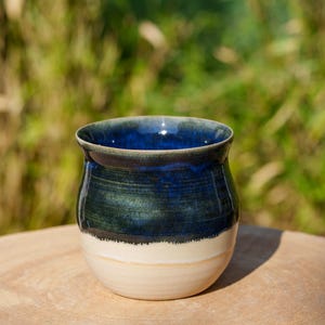 Puede incluir: Una taza de cerámica azul y blanca con forma redondeada. La taza tiene un esmalte azul oscuro con un borde blanco y una base blanca.