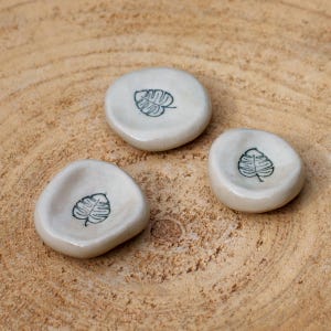 Peut inclure: Trois petits plats en céramique blanche avec un bord surélevé. Chaque plat présente un dessin simple au trait noir d'une feuille de monstera.