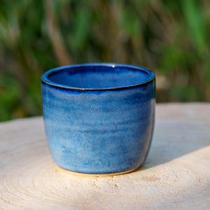 May include: A small, handmade ceramic cup with a deep blue glaze. The cup has a rounded shape and a smooth, textured surface. The interior is a darker shade of blue, and the rim is a darker blue. The cup is sitting on a light-coloured wooden surface.