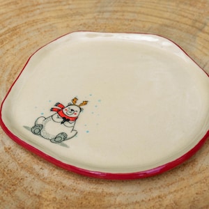 Peut inclure: Une assiette en céramique de couleur crème avec une forme arrondie irrégulière et un fin bord peint en rouge. L'assiette présente une petite illustration d'un ours blanc portant une écharpe rouge et des bois de renne marron, entouré de points bleus.