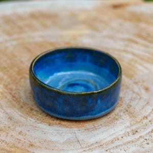 Op de afbeelding: Een kleine, ronde keramische kom met een diepblauwe glazuur. De kom heeft een licht onregelmatige rand en een donkerder blauw interieur. De buitenkant heeft een donkergroene rand. De kom staat op een lichtbruin, getextureerd oppervlak.