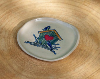 Puede incluir: Un pequeño plato de cerámica verde claro con un diseño de casita de pájaros pintado a mano. La casita tiene un corazón rojo, pájaros azules y detalles florales azules. El plato tiene forma irregular y está sobre una superficie de madera.