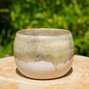 Peut inclure: Un bol en céramique fait à la main, de forme arrondie. Le bol présente un dégradé de couleurs, passant d'une base blanc crème à des nuances de vert, de bleu et de beige en haut. Le bol est posé sur une surface en bois.