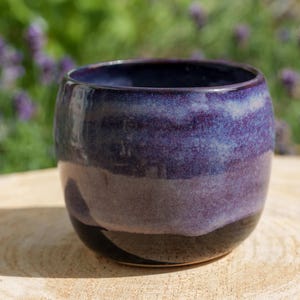 Peut inclure: Une tasse en céramique arrondie dotée d'un vernis brillant avec un dégradé de violet profond sur le dessus, passant par un bleu lavande plus clair au centre, et se terminant par une base noire unie. Le récipient présente un bord lisse et incurvé.