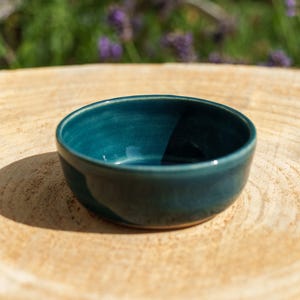Könnte beinhalten: Eine kleine, runde Keramikschale mit einer glatten, glänzenden Oberfläche in einem tiefen Blaugrün. Die Schale hat ein schlichtes, offenes Design mit leicht geschwungenen Wänden und einem flachen Boden, das eine gleichmäßige, einfarbige Glasur zeigt.