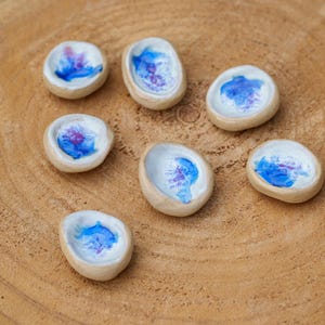 Pode incluir: Sete pequenos pratos de cerâmica feitos à mão com exterior bege natural e interior branco. Cada prato apresenta um design abstrato único em tons de azul e roxo. Os pratos estão dispostos sobre uma superfície de madeira castanha clara.