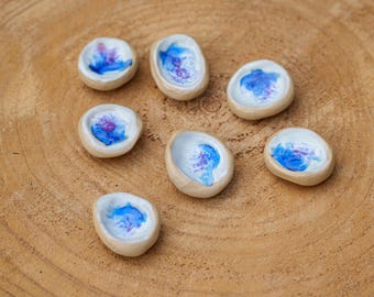 May include: Seven small, handmade ceramic dishes with a natural beige exterior and a white interior. Each dish features a unique abstract design in shades of blue and purple. The dishes are arranged on a light brown wooden surface.