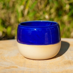 Peut inclure: Une tasse en céramique faite à la main, avec une moitié supérieure bleu cobalt vif et une moitié inférieure beige crème. La tasse est ronde et posée sur une surface en bois, avec un arrière-plan vert flou.