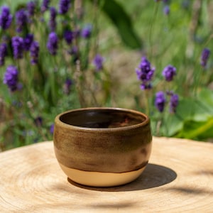 May include: A handmade ceramic bowl with a brown glazed top and a cream-colored base. The bowl is set on a wooden surface, with a blurred background of purple flowers and green foliage.