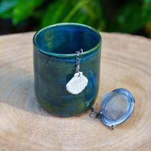Pode incluir: Uma chávena de chá de cerâmica feita à mão com esmalte verde e azul. Um infusor de chá de aço inoxidável e um pingente decorativo pendem da borda da chávena. A chávena está sobre uma superfície de madeira.